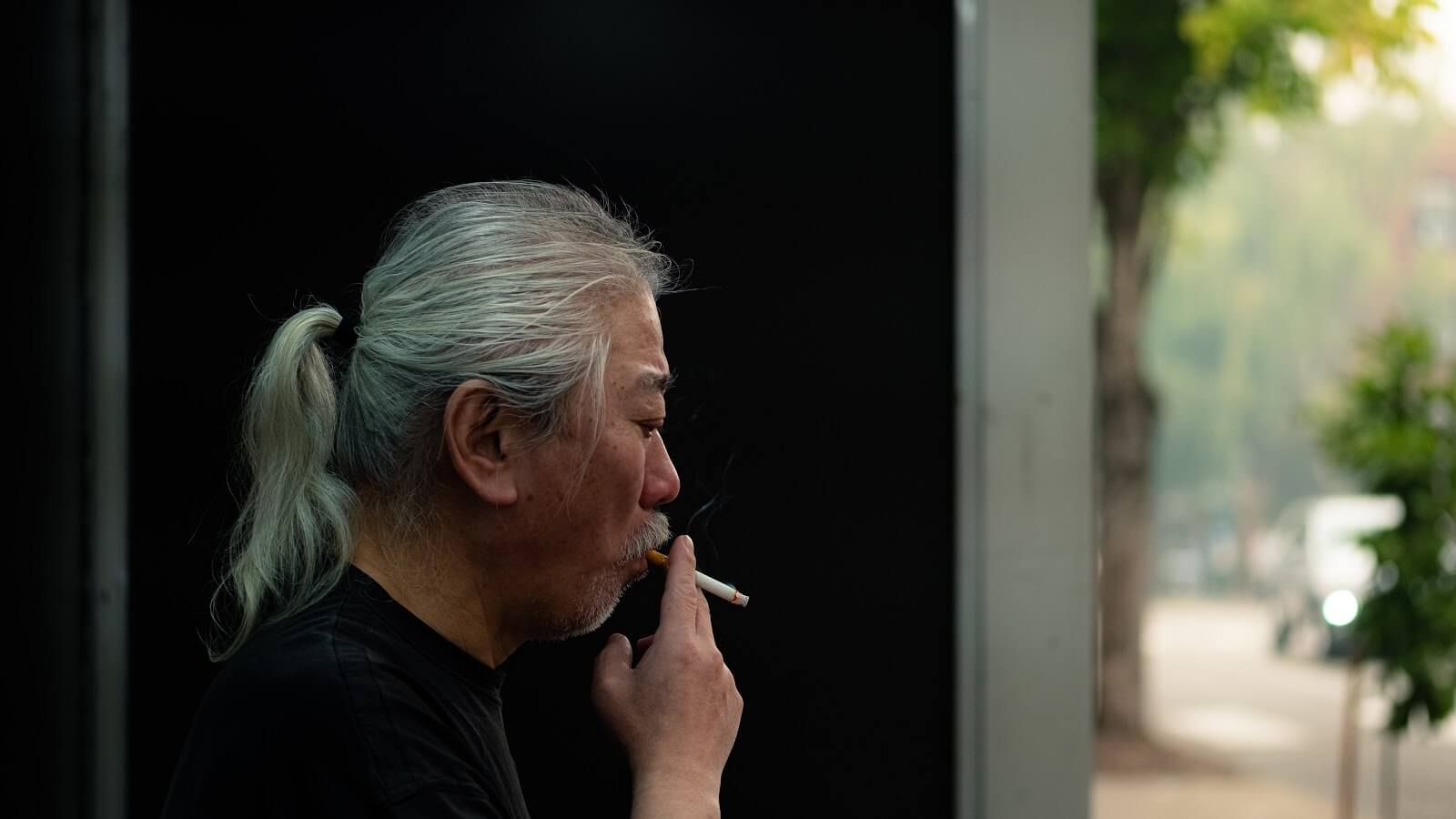 a man with white hair tied back in a ponytail smoking a cigarette